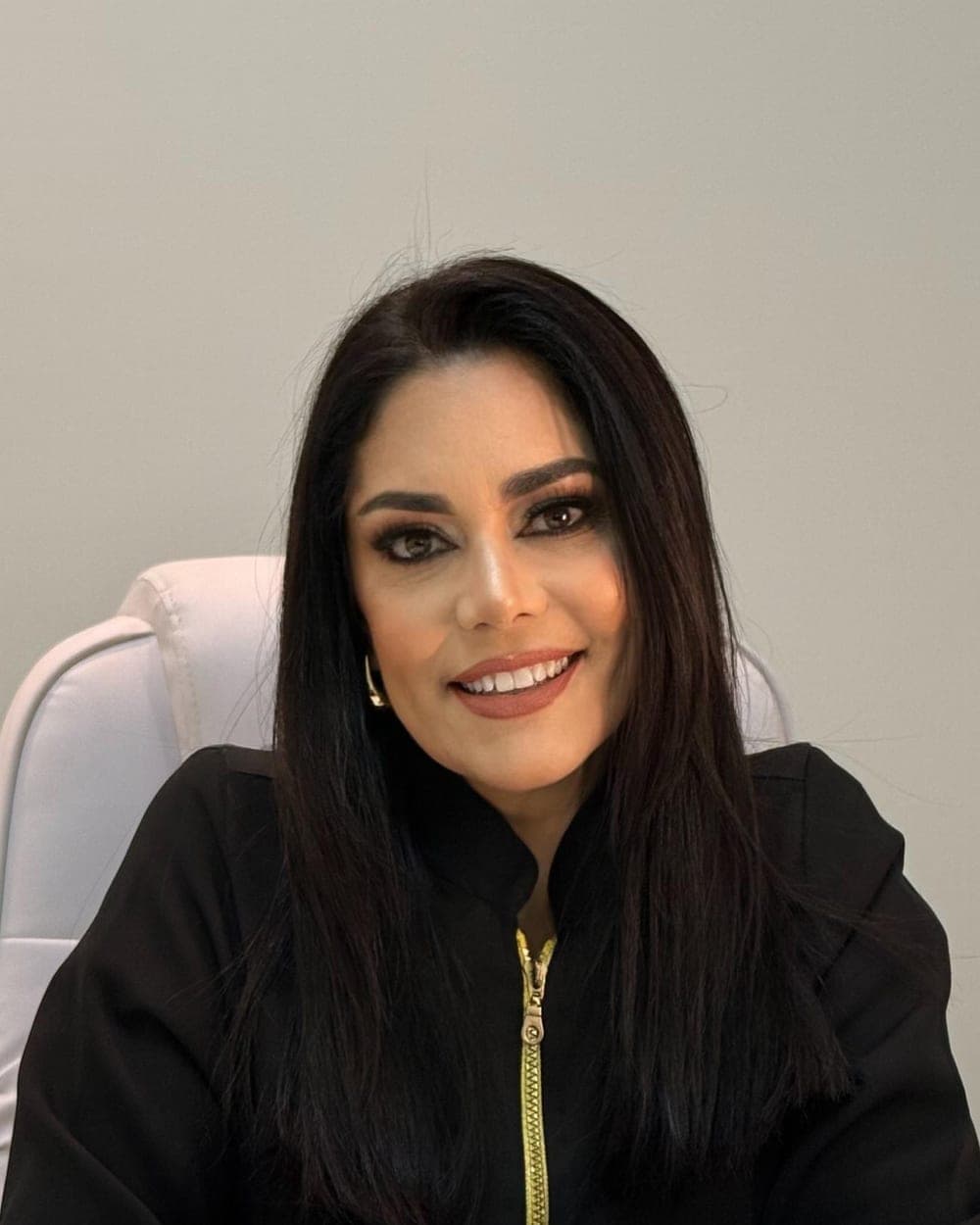 A Dra. Luciana Lemes sorrindo, de jaleco preto, sentada no consultório